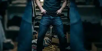 A person struggles with ill-fitting denim jeans amidst scattered clothing, symbolizing outdated sizing and shopping frustration.